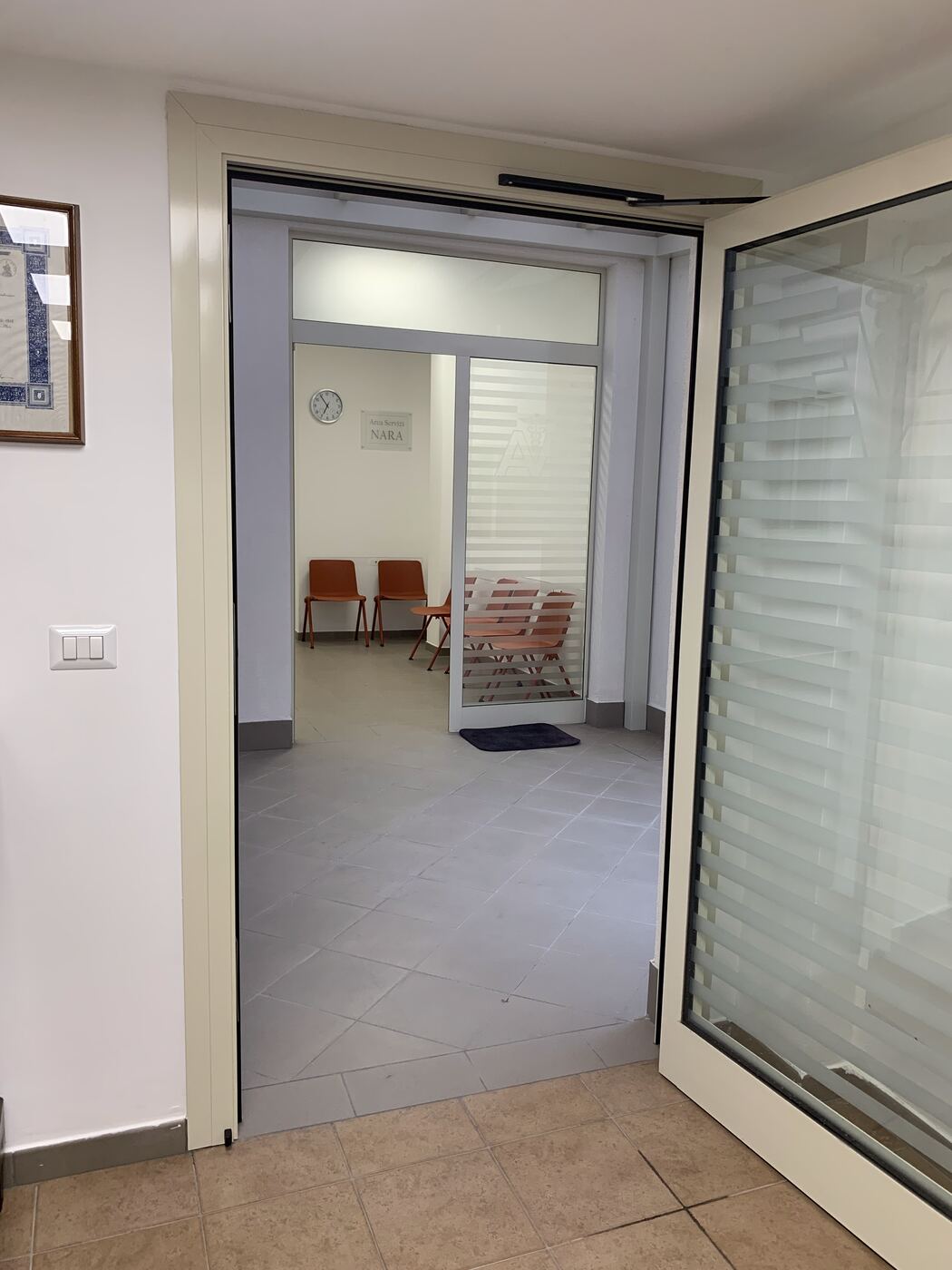 La porta a vetri aperta sull’area servizi della farmacia, con la sala d’attesa sul fondo